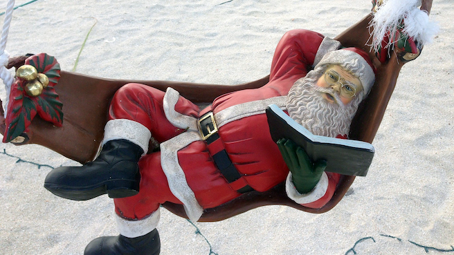 Santa Claus doll laying on a hammock and reading a book by the beach.